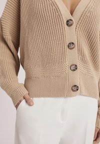 Beżowy sweter z dzianiny z głębokim dekoltem w kształcie litery V i czterema ciemnobrązowymi guzikami. Posiada fakturowane wzory i ściągacze przy mankietach, nałożony na białe spodnie.