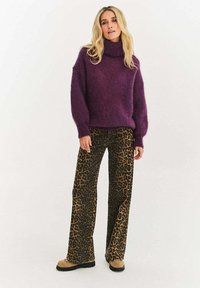 Fialový pletený sveter s vysokým golierom, doplnený hnedými nohavicami s leopardím vzorom. Oblečenie má voľný strih a textúrované materiály.