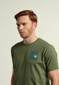 Camiseta de algodón verde con un estampado gráfico que incluye las palabras "GAME SET MATCH" y un icono de bola de tenis en azul y amarillo. Mangas cortas.
