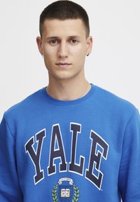 Felpa blu con la scritta "YALE" in lettere bianche e blu navy. Presenta uno stemma al centro, realizzata in morbido materiale misto cotone.