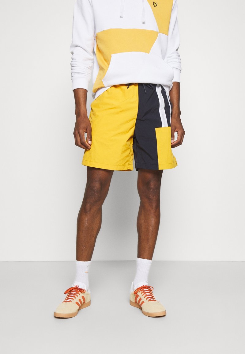 Costumi da bagno gialli e blu navy con tasca laterale e striscia bianca; abbinati a una felpa con cappuccio color block grigia e gialla e scarpe da ginnastica beige.