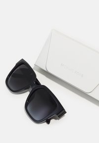 Occhiali da sole neri oversize con lenti in gradiente, montatura in plastica liscia, affiancati a un astuccio per occhiali bianco e testurizzato, inciso con "Michael Kors".