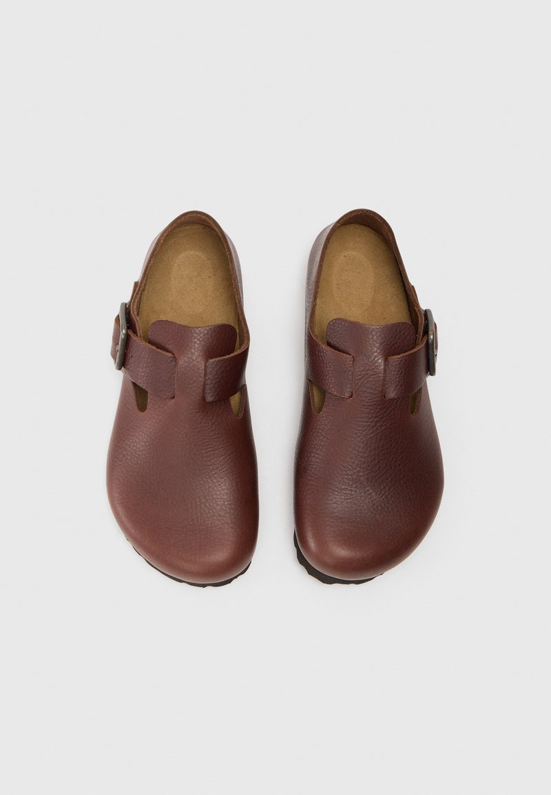 Zuecos de cuero marrón con un acabado suave, que presentan detalles recortados y una hebilla lateral. La suela es texturizada para un mejor agarre.
