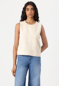 Top senza maniche di colore crema con dettagli in perle in rilievo, scollatura rotonda e orlo dritto. Abbinato a pantaloni in denim azzurri.