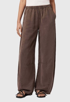 Luźne brązowe spodnie z elastycznym pasem, przednie kieszenie, noszone z czarnymi sandałami z odkrytymi palcami na osobie stojącej na jednolitym tle.