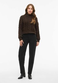 Ciemnobrązowy sweter w dżersejowy splot z wysokim dekoltem, w połączeniu z czarnymi spodniami o prostym kroju i czarnymi botkami na kostkę. Gładka tekstura i dopasowany krój.