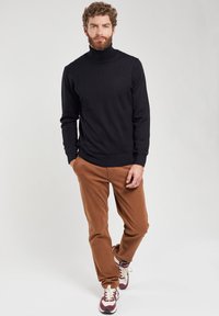 Maglione a collo alto nero in un tessuto morbido, abbinato a pantaloni marroni. Sneakers bianche e bordeaux completano l'outfit. Design semplice e moderno.