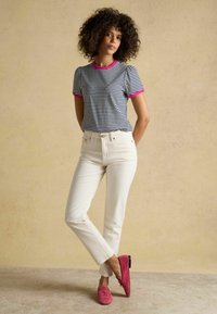 Žena s kučeravými vlasmi oblečená v čierno-bielom pruhovanom tričku s ružovým lemovaním, biele nohavice a ružové mokasíny, stojaca na béžovom pozadí.