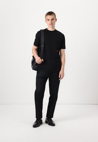 Svart kortermet T-skjorte med et lite rødt design, kombinert med svarte bukser og sko. Modellen holder en svart veske og står foran en hvit bakgrunn.