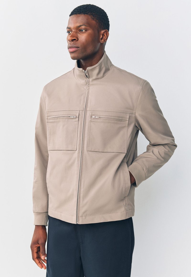 Jeune homme en veste beige zippée avec poches poitrine, debout avec une main dans la poche, regardant légèrement sur le côté devant un fond uni.
