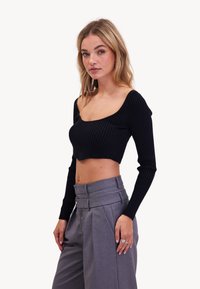 Zwarte geribbelde crop top met lange mouwen; heeft een brede ronde halslijn. Gepaard met hoogoplopende grijze geplooide broeken.