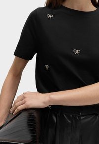 Černé bavlněné tričko s malými krystalovými mašličkami, krátkými rukávy a kulatým výstřihem. Model drží černou lakovanou kabelku.