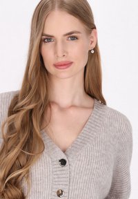 Světle šedý žebrovaný cardigan s výstřihem do V, dvěma černými knoflíky a měkkou texturou. Dlouhé, vlnité vlasy rámují obličej.