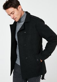 Cappotto di lana nero con design doppiopetto, grandi bottoni e tasche laterali; abbinato a un dolcevita grigio chiaro sottostante.