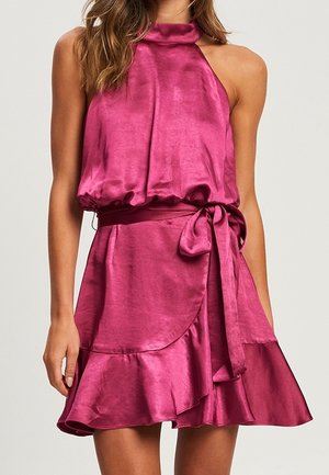 Vestido sin mangas de satén magenta con cuello halter, lazo en la cintura y falda asimétrica con volantes, llevado por una persona de pie.