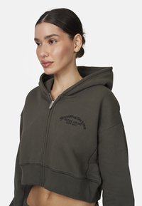 Afgebroken hoodie in donkergroen met een ritssluiting aan de voorkant. Kenmerkt zich door geborduurde tekst, contrasterende stiksels en een zachte stofstructuur.