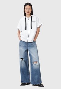Sudadera blanca con cremallera y cordones negros, acompañada de jeans anchos de mezclilla azul desgastados, con dobladillos deshilachados y grandes desgarros.