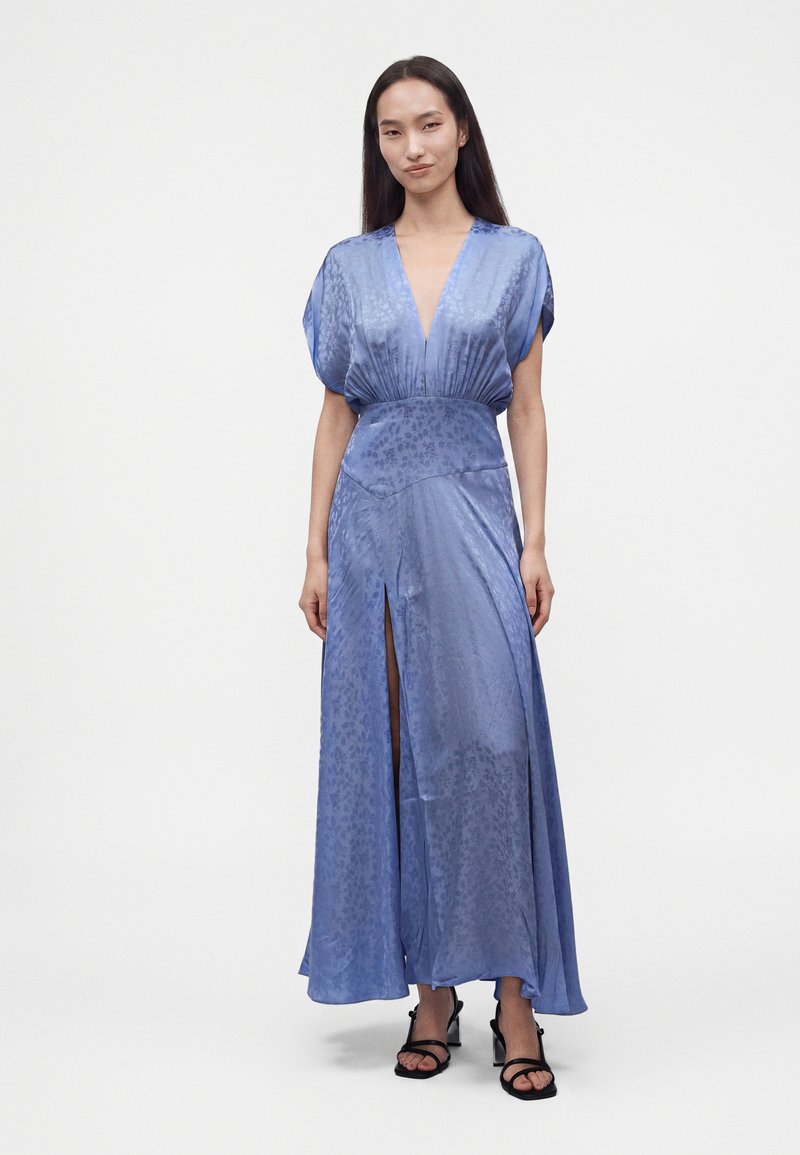 Femme portant une robe longue en satin bleu avec un profond décolleté en V et une fente devant, assortie à des sandales à talons noires à lanières, sur un fond uni.