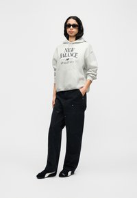 Γκρι φούτερ με κουκούλα με στάμπα "NEW BALANCE ATHLETICS", συνδυασμένο με φαρδιά μαύρα παντελόνια και μαύρα παπούτσια με λευκές λεπτομέρειες. Φοράει μαύρα γυαλιά ηλίου.