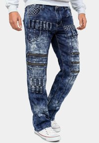 Jeans in denim blu scuro con una texture sbiadita. Presentano molteplici tasche, cerniere e cuciture decorative lungo i lati.