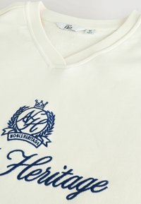 Tricou alb cu mânecă scurtă și guler în V, dintr-un material cu caneluri, având un logo și text brodat în bleumarin pe față.
