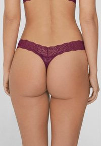 String en dentelle de couleur bordeaux profond avec un motif floral. Présente une taille scallopée, un minimum de couverture et une texture en dentelle élaborée.