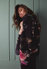 Kurtka z polaru w kwiatowy wzór na ciemnym materiale, z wysokim kołnierzem i luźnym krojem, prezentująca duże różowe i zielone kwiaty na czarnym tle.