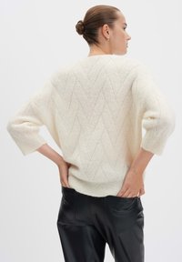 Frau mit einem Dutt, die einen strukturierten cremefarbenen Strickpullover und schwarze Lederhosen trägt, mit den Händen in den Hosentaschen, die von hinten blickt.
