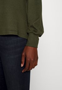 Olivgrön stickad tröja med lös passform, med ribbad manschett och avslut. Buren med mörka jeans i denim, vilket visar en närbild av ärmen och handen.