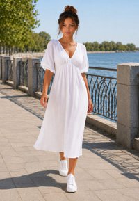 Donna in abito midi bianco e sneakers bianche che cammina lungo una passeggiata sul lungofiume con alberi e ringhiere sullo sfondo in una giornata di sole.
