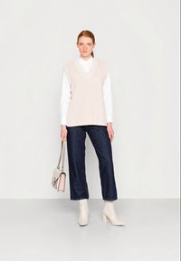 Woman wearing beige knit vest, white shirt, dark jeans, white ankle boots, holding a beige handbag with a chain strap against a plain background.