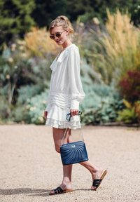 Bílé šaty s volánkovým lemem, modrá prošívaná kabelka s řetízkem a černé sandály na klínu; umístěné v zahradě se zeleným pozadím.