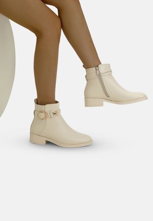 Bottines beiges en matériau synthétique lisse, avec une fermeture éclair sur le côté et une boucle décorative sur la sangle de la cheville. Talon plat.