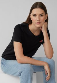 Zwarte katoenen T-shirt met korte mouwen en ronde hals, met een kleine borduursel van een rode en witte watermeloenschijf op de borst.