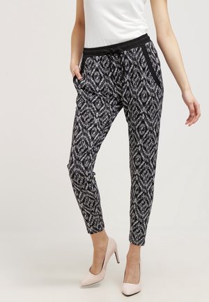 Nainen, joka pukeutuu mustavalkoisiin abstraktin timanttikuosin juoksuhousuihin, joissa on musta vyötärönauha, ja valkoisiin korkoihin, seisoo käsi taskussa.