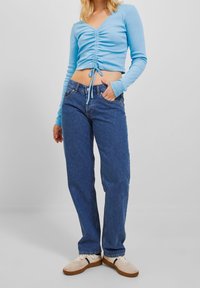 Helles blaues Langarmoberteil mit Raffung und Bindebändern an der Taille, kombiniert mit hochgeschnittenen dunkelblauen Jeans und beigen Sneakers.