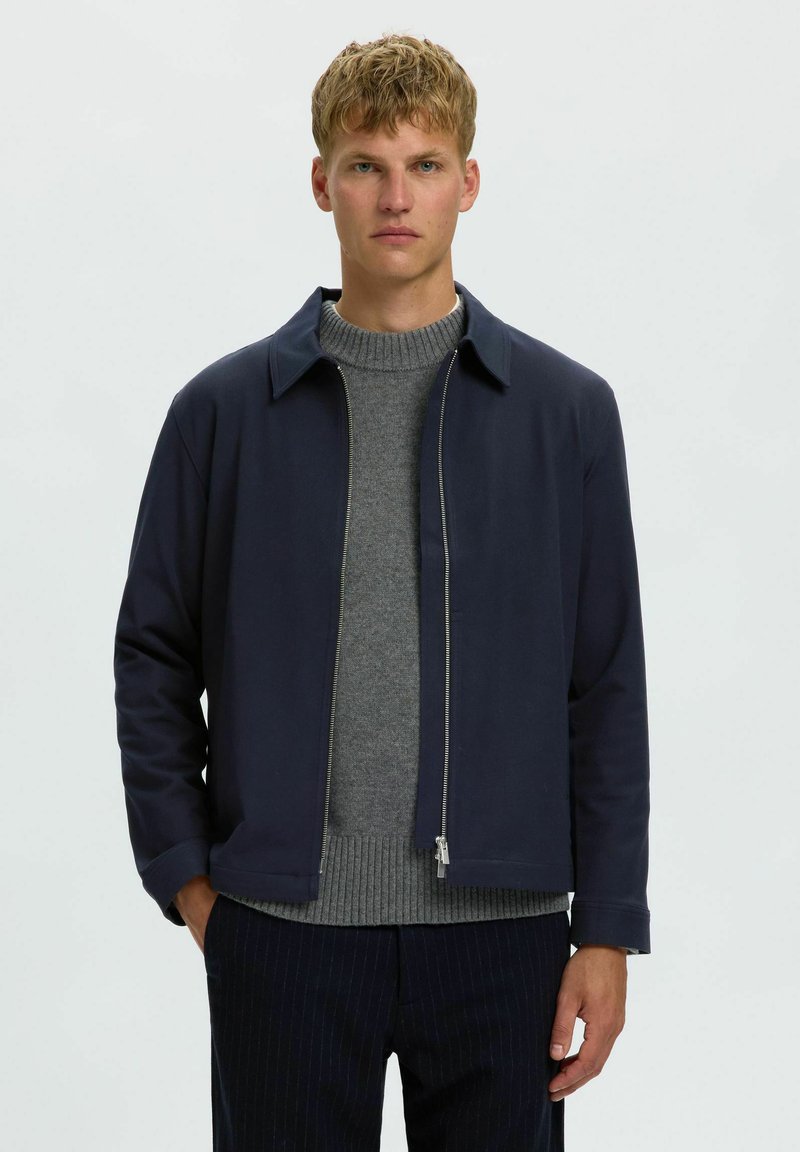 Homme blond portant une veste bleu marine zippée sur un pull en maille gris et un pantalon sombre à fines rayures, debout devant un fond clair uni.