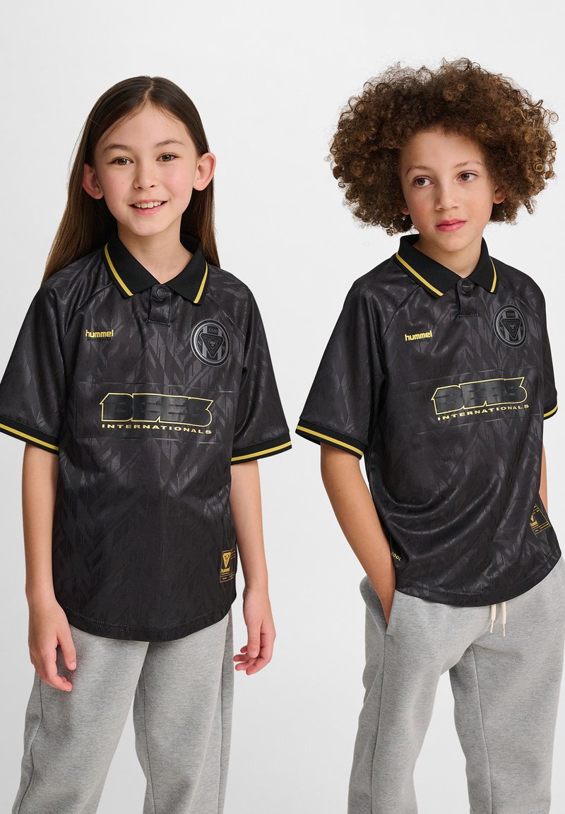 Svart kortärmad tröja med en texturerad design, gula accenter och krage. Har två logotyper och en avslappnad passform, tillsammans med grå byxor.