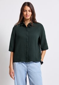 Camicetta button-up verde scuro, con texture e maniche corte, dotata di colletto e una vestibilità rilassata, abbinata a jeans azzurri chiari e larghi.