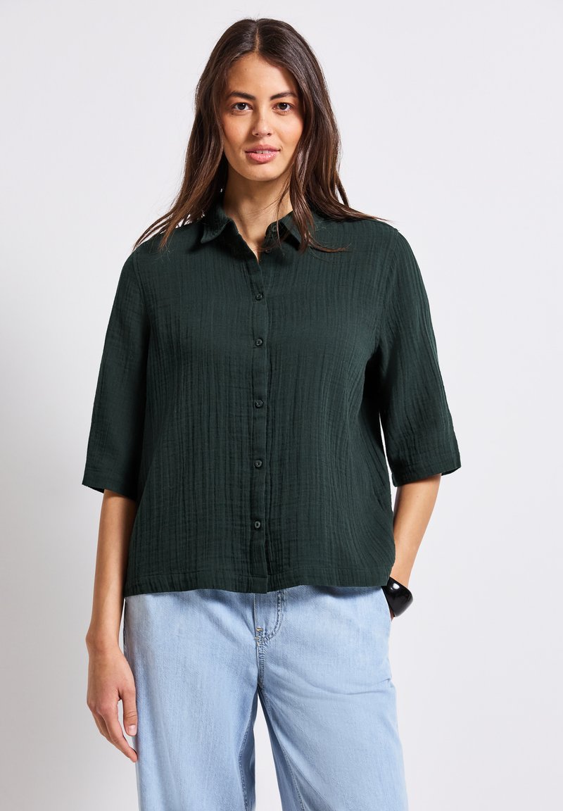 Camicetta button-up verde scuro, con texture e maniche corte, dotata di colletto e una vestibilità rilassata, abbinata a jeans azzurri chiari e larghi.