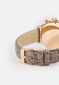 Rožinio aukso laikrodis su tekstūruotu šviesiai pilku odiniu dirželiu, turintis metalinį užsegimą, išgraviruotą žodžiu "SWAROVSKI," demonstruojantis subtilų blizgesį ir detales.