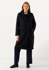 Námorně modrý kabát ze směsi vlny s dvojitým zapínáním a velkými knoflíky, klopovým límcem a bočními kapsami, v kompozici s námořnickými kalhotami a černými botami.