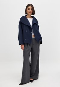 Tume sinise värvi kahekordse nööbiga mantel suure kraega, millele on kombineeritud hallid laiad püksid; mõlemad on valmistatud struktuursest kangast. Mustad terava turjakujulise ninaga kingad.