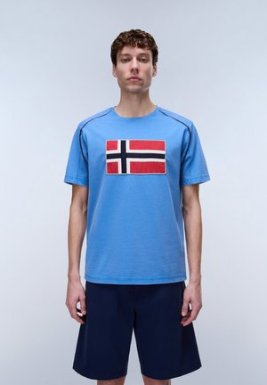 Ung mand med kort krøllet hår iført en blå t-shirt med et norsk flagmærke og marineblå shorts, stående foran en ensfarvet baggrund.