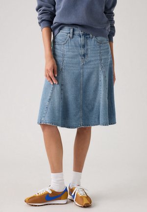 Porte une jupe en jean bleue longueur genou, des chaussettes blanches et des baskets marron avec un swoosh Nike bleu, associées à un haut bleu à manches longues.