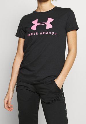 Osoba w czarnej koszulce Under Armour z różowym logo i napisem, w czarnych spodniach, z jedną ręką w kieszeni, na jednolitym tle.