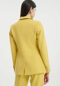 Blazer giallo su misura con un design strutturato, apertura singola sul retro e dettagli con bottoni sui polsini. Tessuto con una texture liscia.