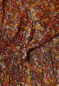 Tissu avec de petites fleurs jaunes, orange et blanches sur un fond marron, présentant une manche froncée avec un poignet élastique et une texture plissée.