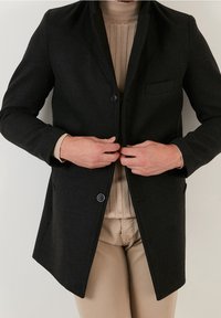 Cappotto nero in misto lana con chiusura a un bottone, rever a incavo e taschino sul petto. Indossato sopra un maglione beige testurizzato.