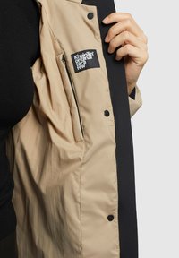 Beige jakke med sort detaljer indeni, forsynet med et logo-lap og trykknap-lukninger. Glat tekstur med et let udseende.
